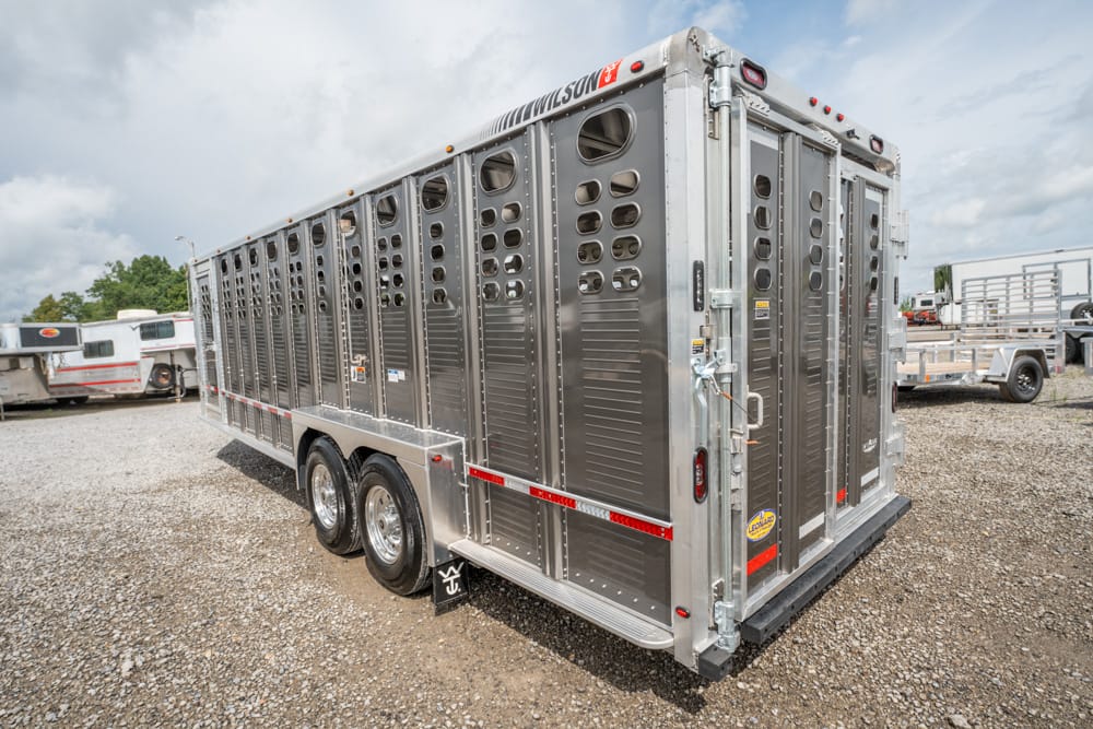 Metal livestock trailer with numerous ventilation holes parked on a gravel lot, rear doors closed and dual axles visible.