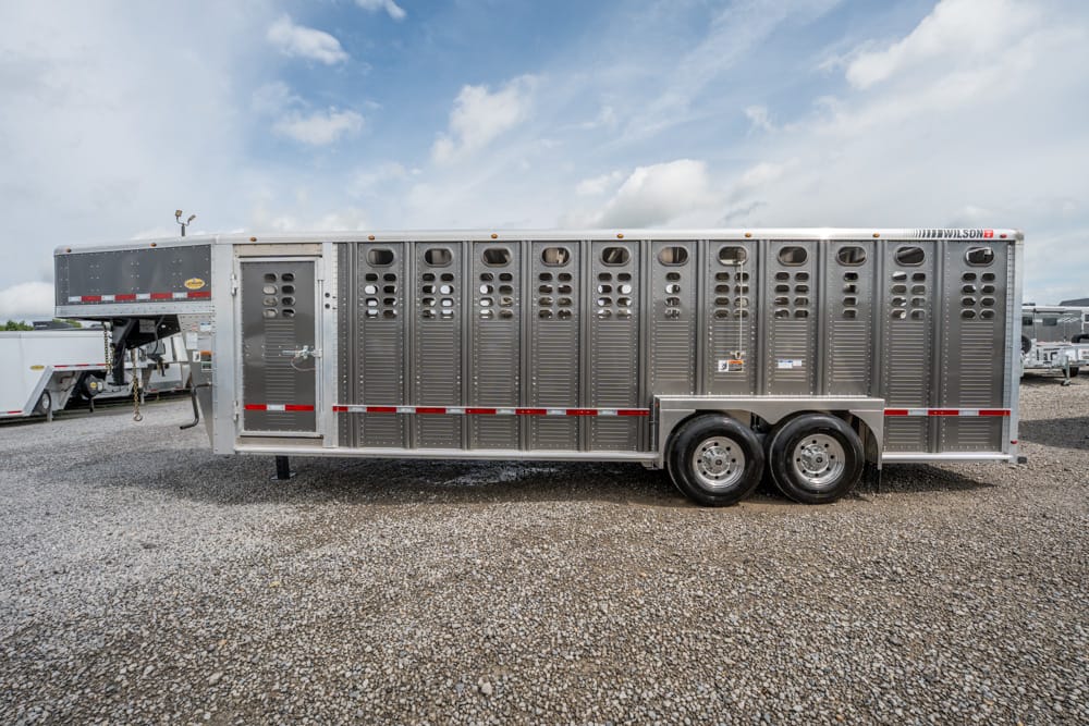 Long aluminum livestock trailer parked on a gravel lot, with ventilation slots along the sides and tandem axles with dual wheels.