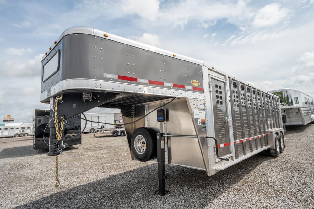Aluminum gooseneck trailer with ventilated stalls parked on a gravel lot, landing gear extended and safety chains visible.
