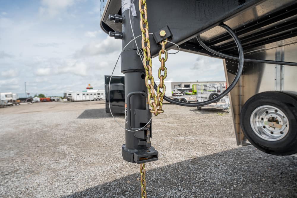 Close-up of a trailer tongue with safety chains, electrical line, and hitch pin, parked on a gravel lot in a trucking yard.