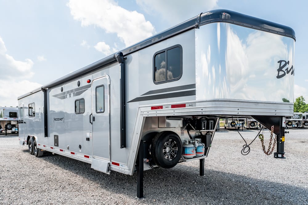 Silver fourth-wheel style RV trailer on a gravel lot, showing the raised front hitch and propane tanks at the hitch area.