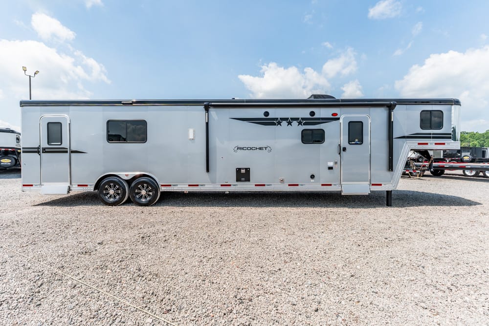 A large gray fifth-wheel RV trailer with two axles parked on a gravel lot under a blue sky, labeled Ricochet with an extended living area.