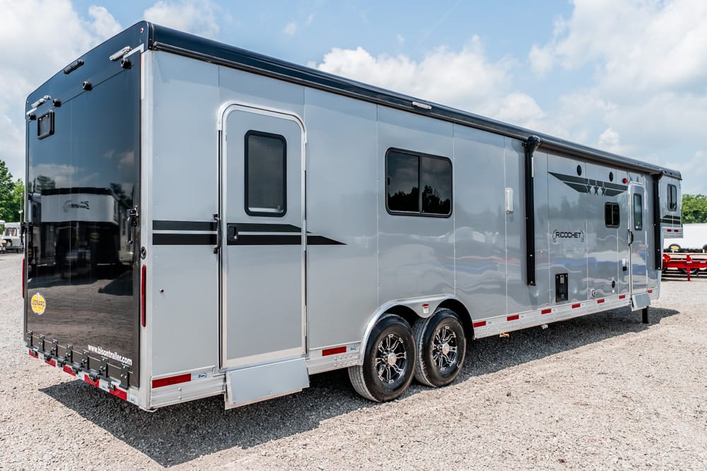 Long silver travel trailer with two axles parked on gravel under a blue, partly cloudy sky.