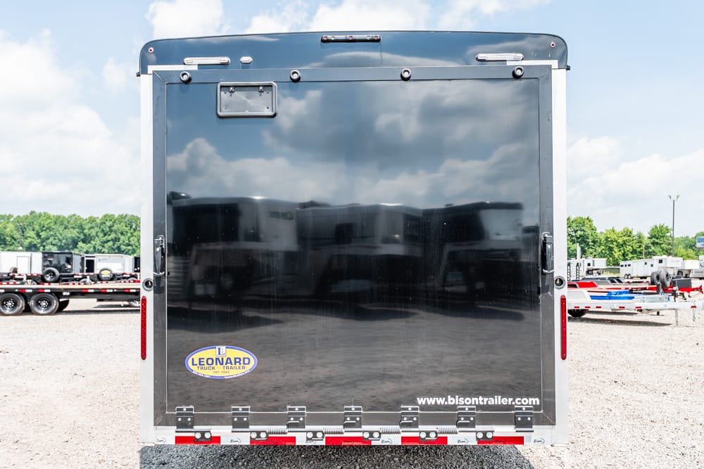 Back of a glossy black trailer with a Leonard logo and the web address www.bisontrailer.com, parked in a gravel lot with other trailers in the background.