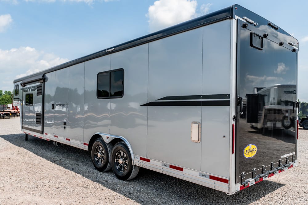 Silver enclosed travel trailer with two axles and a side window, parked on a gravel lot with a black stripe decal along the side.
