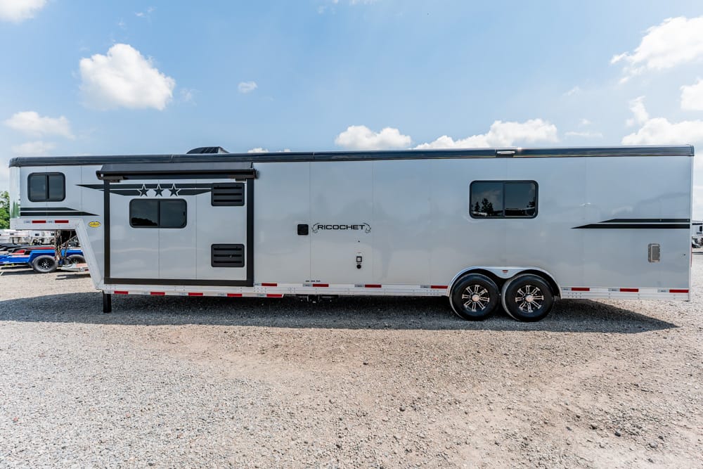 Silver Ricochet travel trailer with a side slide-out, parked on a gravel lot under a blue sky with clouds.