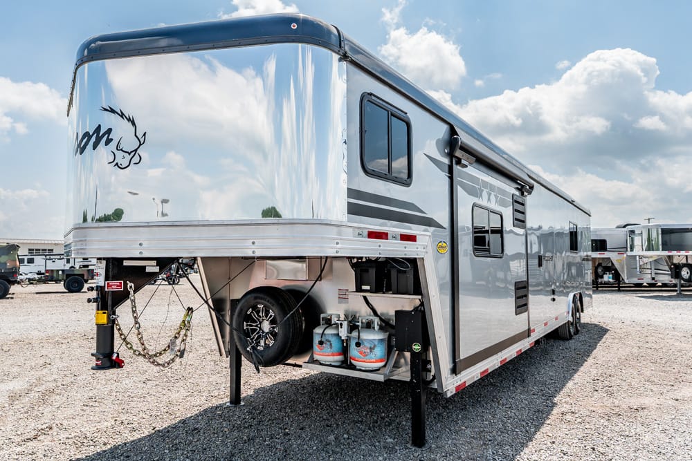 Silver gooseneck travel trailer parked on gravel; front hitch area shows chains, stabilizers, a spare tire, and propane tanks visible beneath the unit.