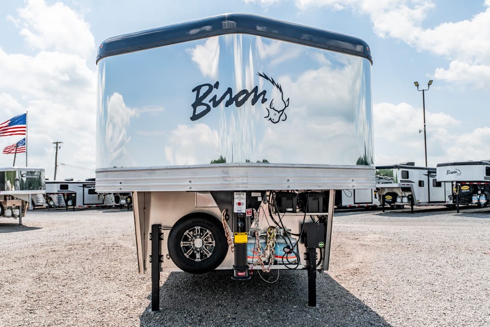 Front view of a chrome Bison trailer with a spare tire and hitch components visible under the raised front, in a gravel lot with other trailers in the background and an American flag nearby.