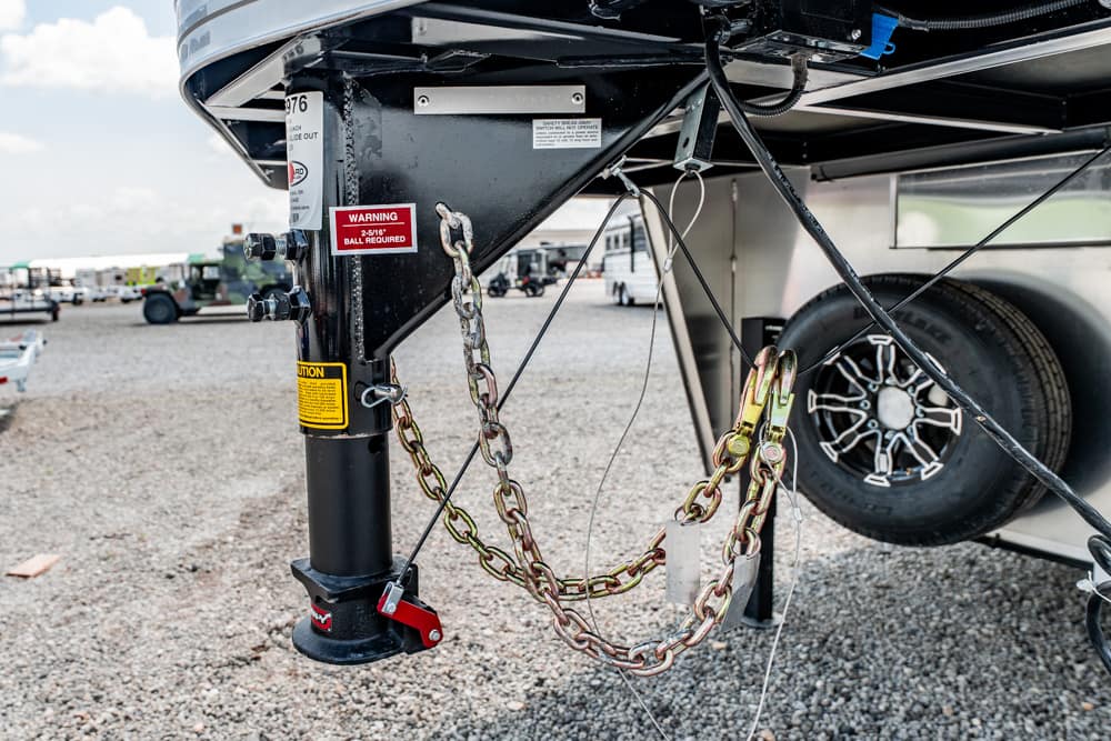 Close-up of a trailer hitch with safety chains and a red pin, tire and gravel yard in the background, warning labels visible.