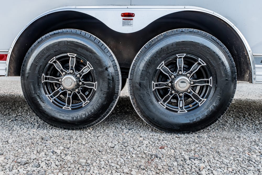 Dual trailer tires with chrome rims on a gravel surface under a silver trailer chassis.
