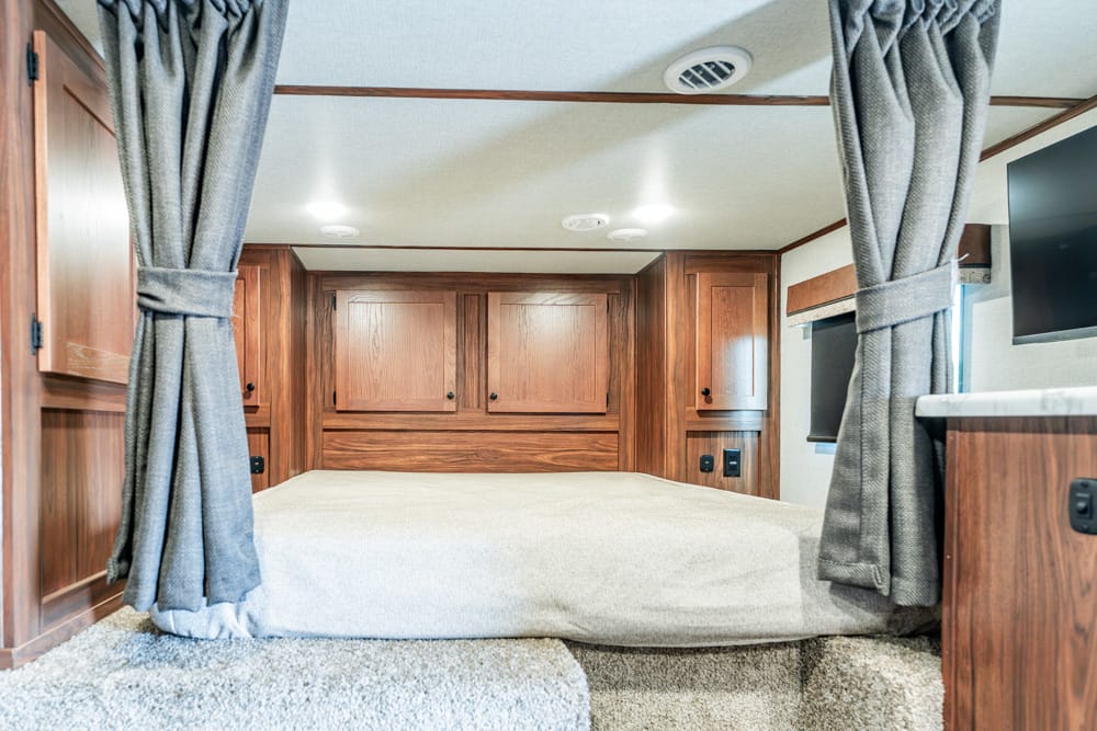 Wooden RV bedroom with a low, gray bed and built-in overhead cabinets; gray curtains frame the sides.