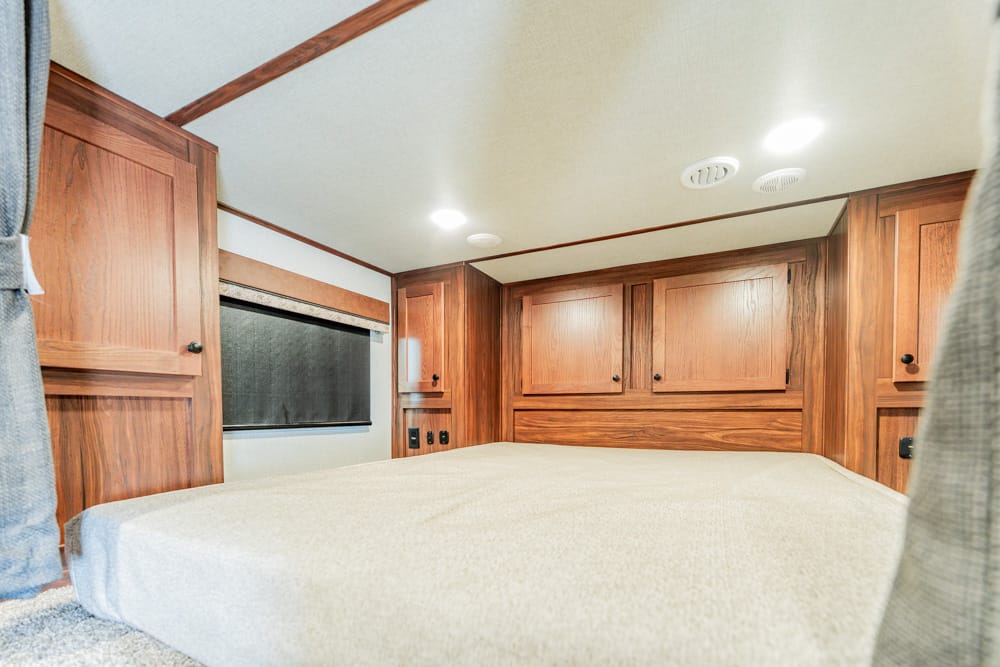 Wood-paneled RV bedroom with a large beige bed, built-in cabinets, and a window with a shade.