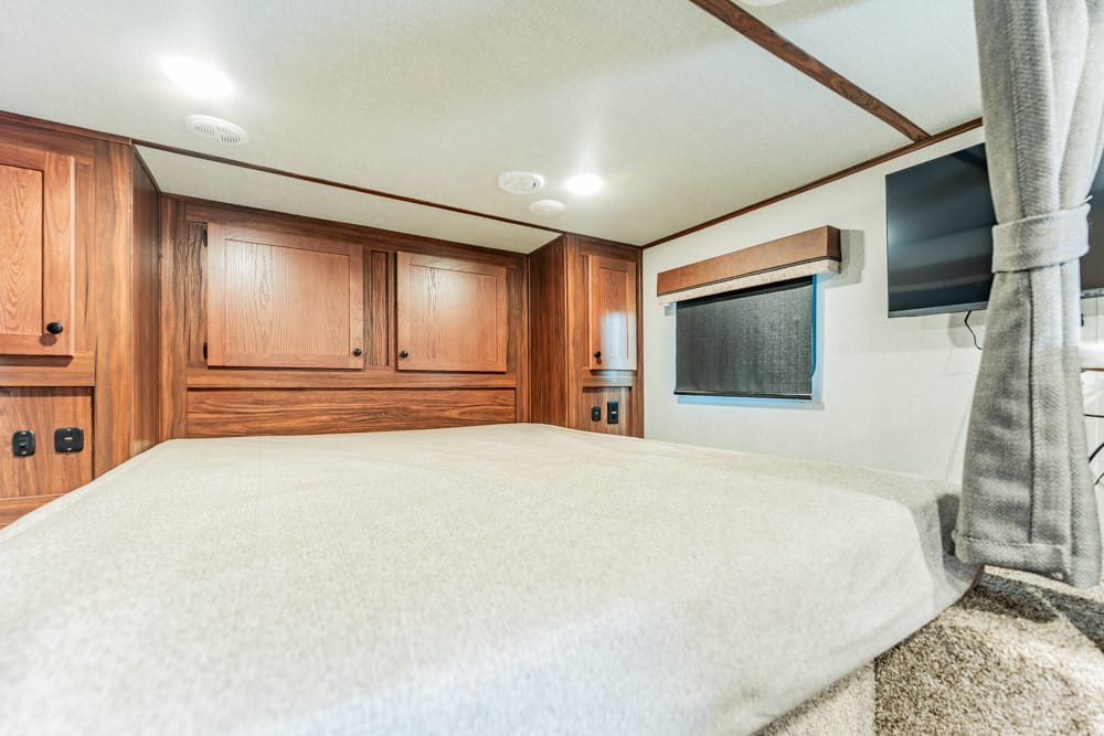 Interior of a camper bedroom with dark wood cabinets above a beige bed, a small window with shade, and a wall-mounted TV.