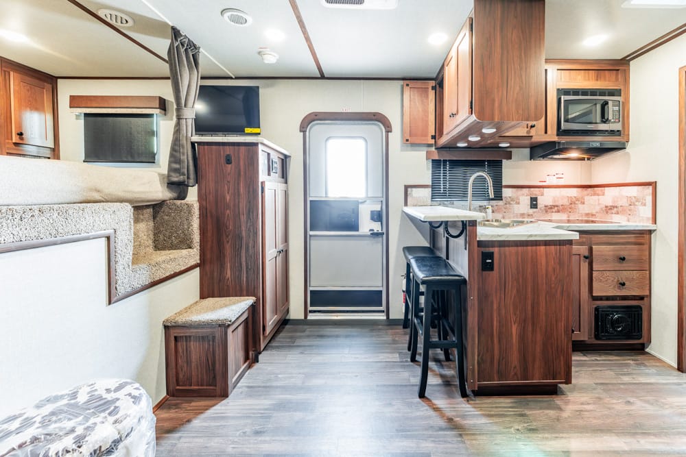 Interior of a travel trailer showing a compact kitchen with wood cabinets, island, sink, and seating, plus a rear entry door.