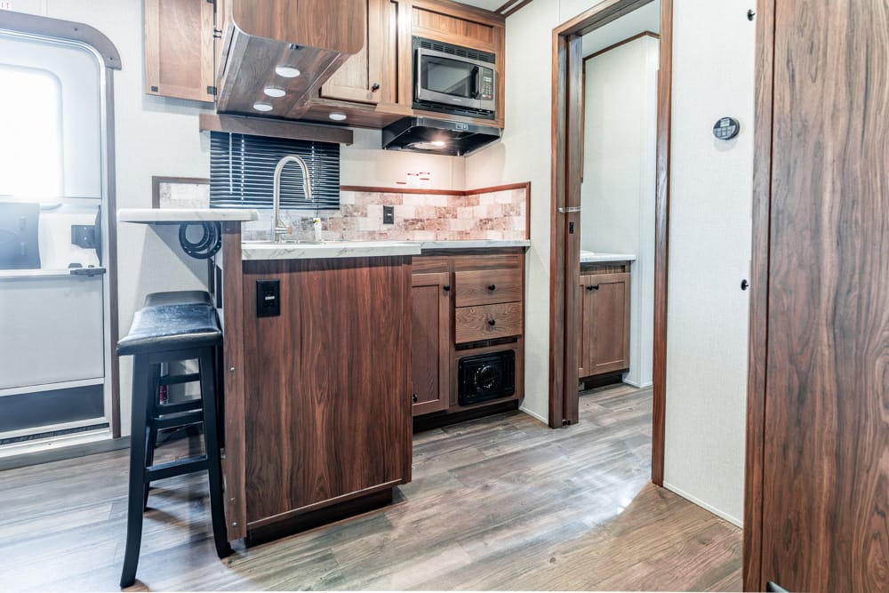 Compact RV kitchen with wood cabinets, sink, stove, and microwave, plus a bar stool at the counter.