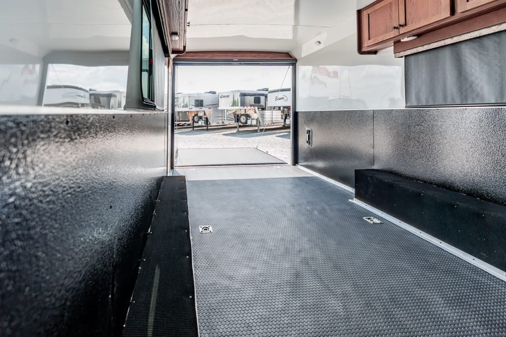 Interior of an empty toy hauler/trailer with textured metal walls, diamond-plate floor, and wooden cabinets above, open to outdoors where RVs are parked.