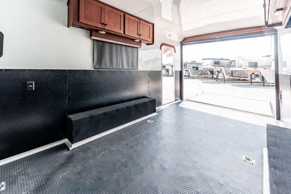 Interior of a trailer showing black padded bench along the wall, wood overhead cabinets, and a textured gray floor; rear door open to a row of parked trailers outside.