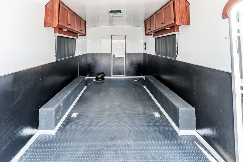 Interior of an empty utility trailer with dark lower walls, bench seating along both sides, and wooden cabinets overhead.