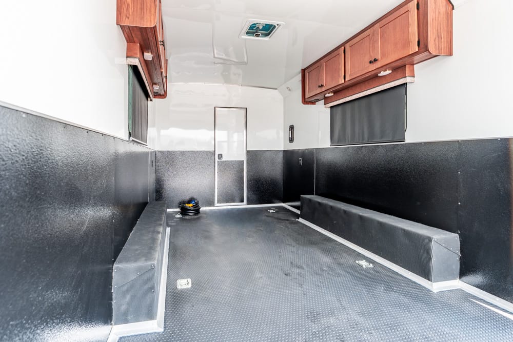 Interior of an empty utility trailer with black walls, diamond-plate floor, and wooden overhead cabinets on top sidewalls.
