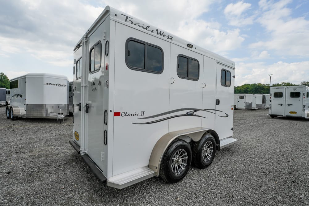 White horse trailer with dual axles in a gravel lot, featuring black swoosh graphics and 'Trails West Classic II' branding near the top edge.