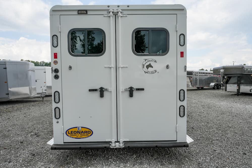 Rear view of a white double horse trailer with two small windows and black latches on a gravel lot, Leonard trailer sticker visible on bottom left.