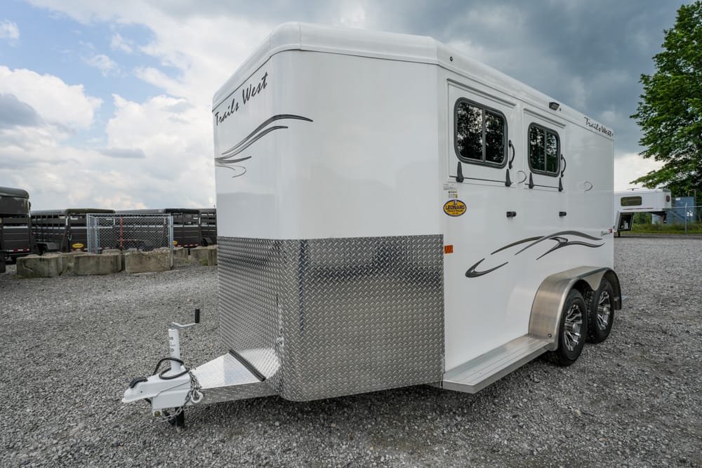 White two‑axle horse trailer with a diamond‑plate front and a hitch on a gravel lot, featuring two windows and decorative gray swoosh graphics on the side.