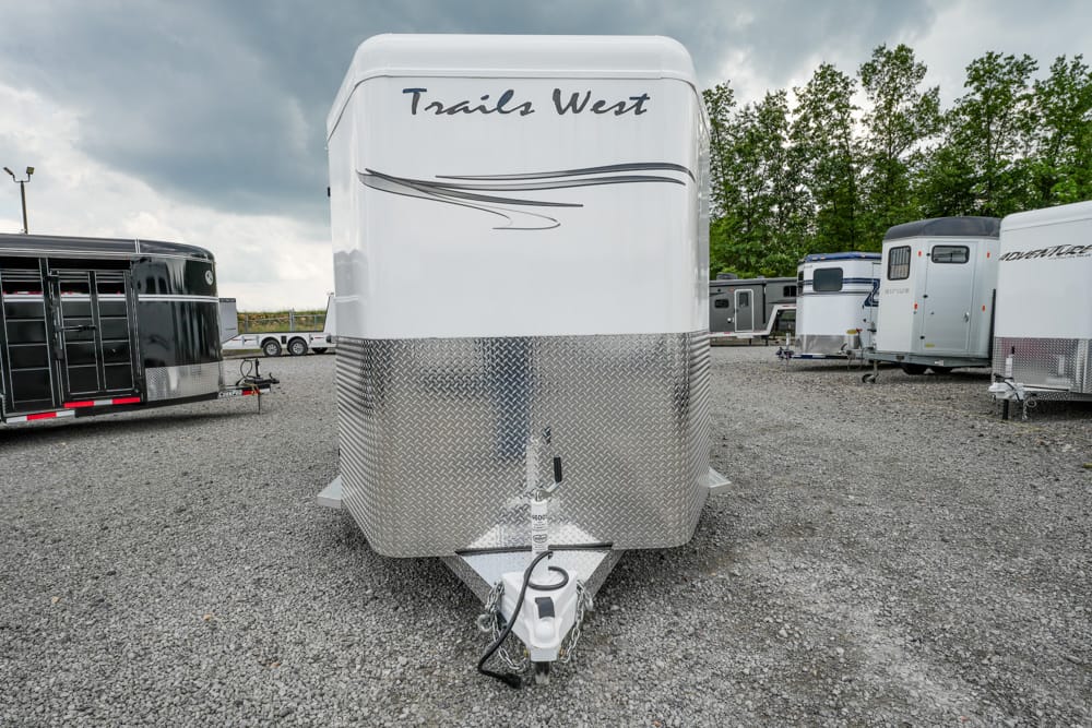 Front view of a white Trails West bumper-hinged horse trailer with a diamond-plate metal front, parked on a gravel lot among other trailers; hitch and safety chain visible at the tongue.