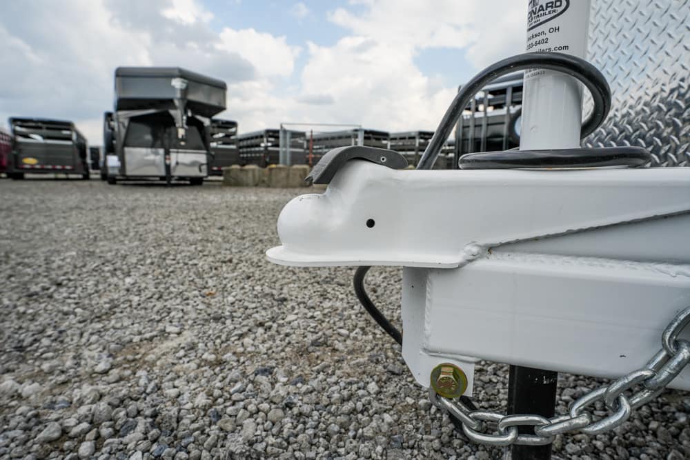 Close-up of a white trailer hitch with safety chains on a gravel lot, with other trailers in the background.