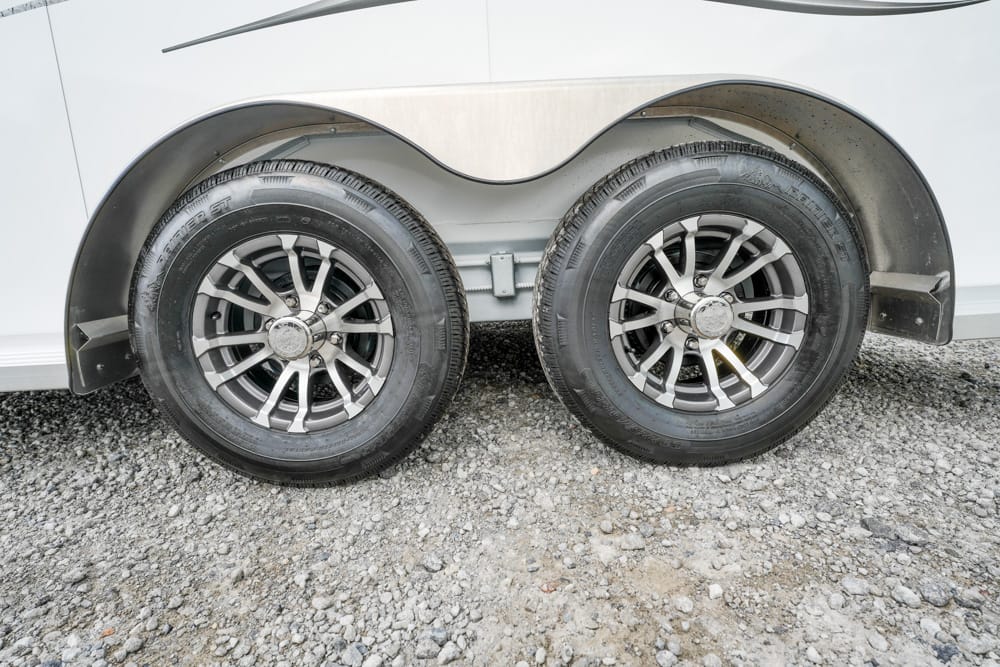 Dual-axle trailer wheels with chrome rims mounted on a gravel surface under a silver trailer.
