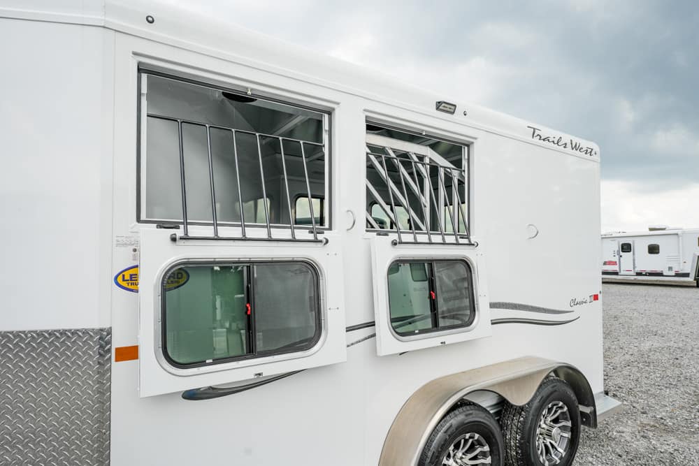 White horse trailer with two front windows open and metal bars, parked on a gravel lot with other trailers in the background, dual-axle wheels visible.