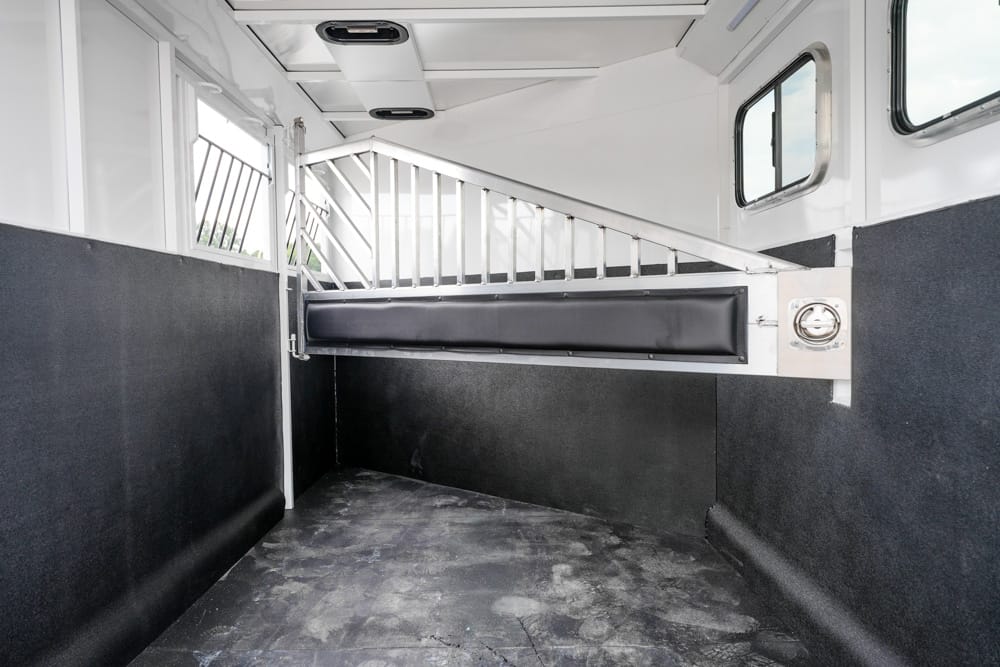 Interior of a horse trailer with a padded wall and a metal divider creating a stall, dark floor, and small windows near the top left and right.