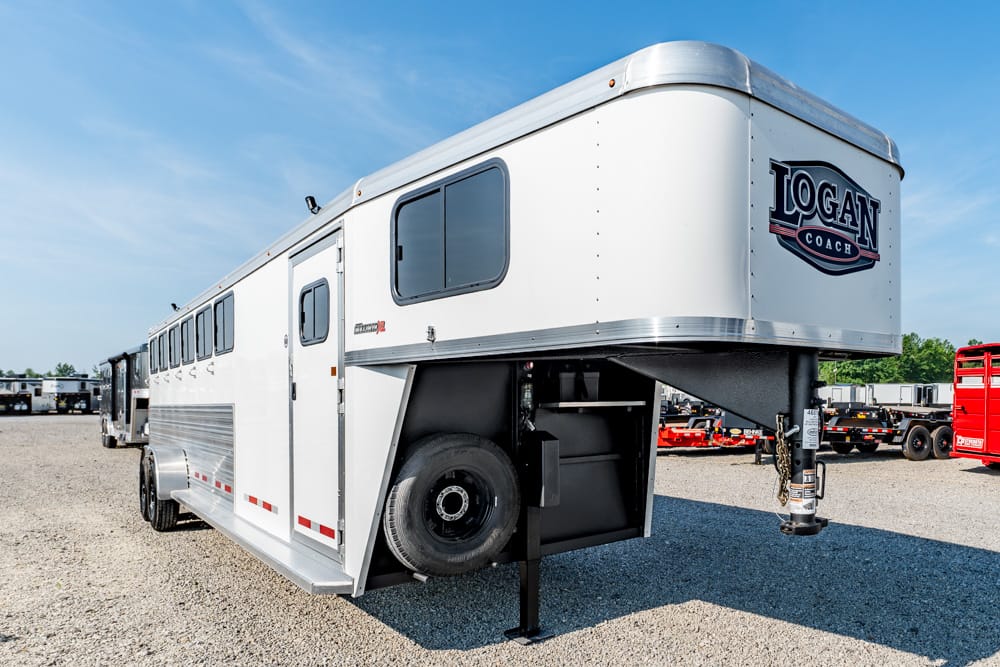 White Logan Coach horse trailer on a gravel lot, with a gooseneck hitch and spare tire mounted on the side while parked.