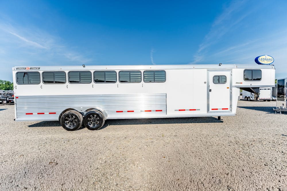 White double-axle horse trailer with windows, parked on a gravel lot under a bright blue sky. Gently curved aluminum siding and red reflective strips are visible.