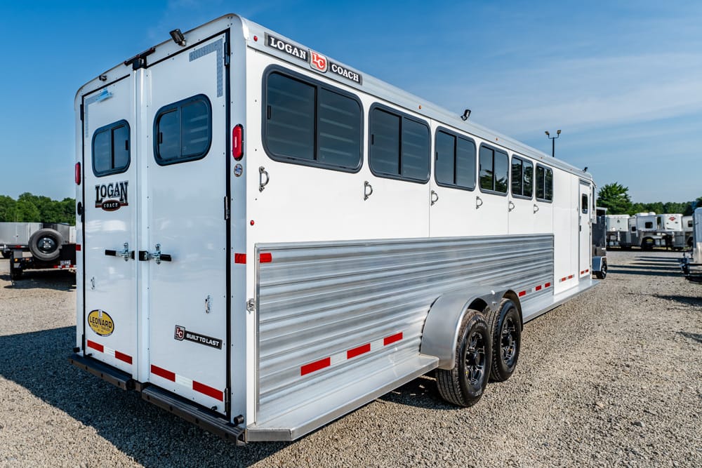 White Logan Coach horse trailer with side windows, parked on a gravel lot under a blue sky.