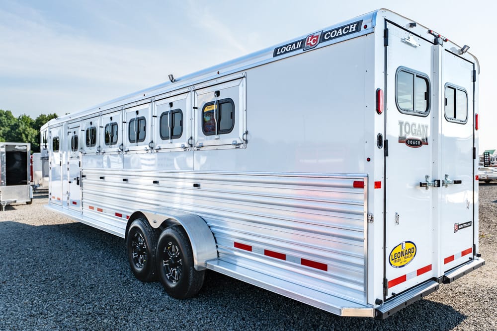 White Logan Coach horse trailer with multiple side windows and dual axles, parked on a gravel lot.
