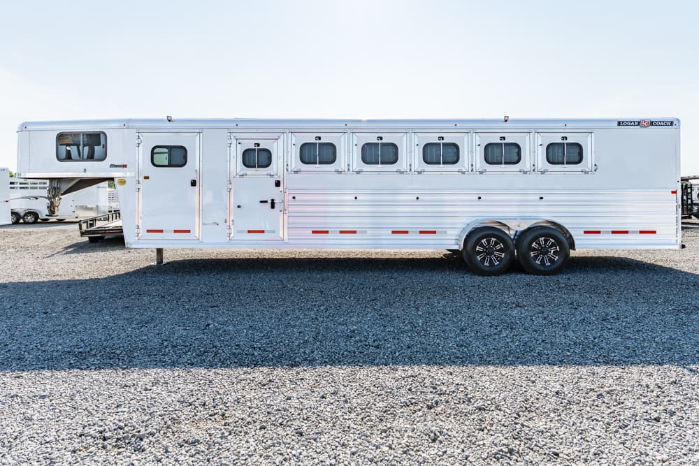 White horse trailer with multiple windows and dual axles parked on a gravel lot on a sunny day.