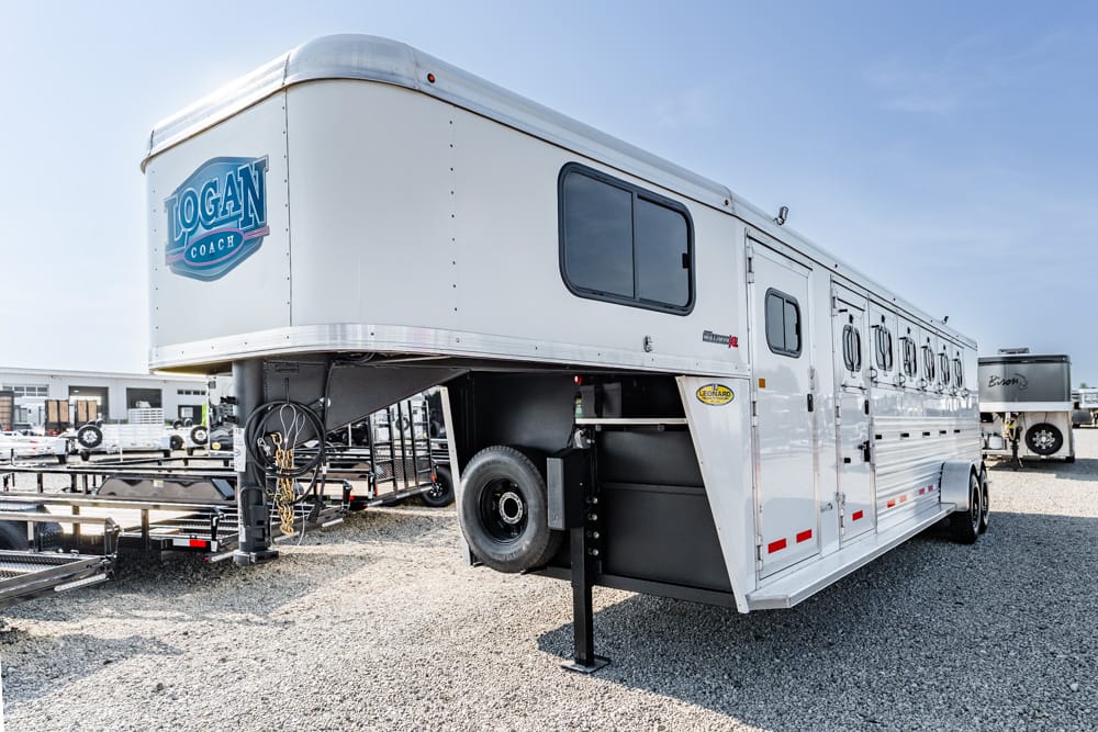 White gooseneck horse trailer with Logan Coach logo, parked on a gravel lot with a raised front section and spare tire visible.