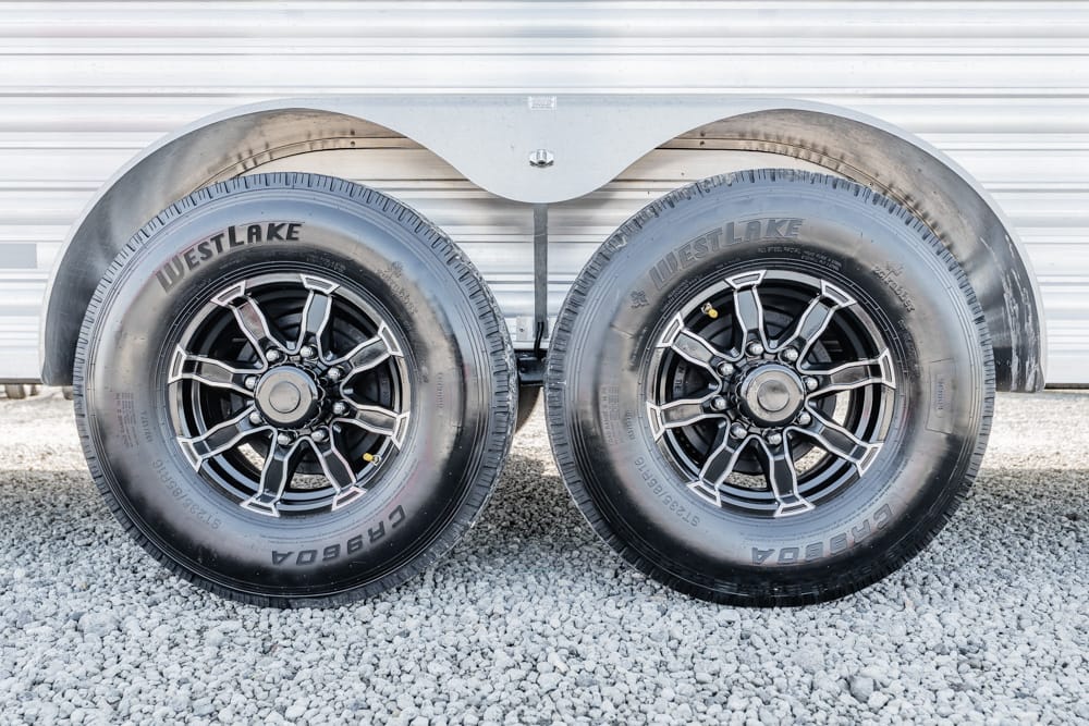 Dual trailer wheels with black and silver spoke rims on a gravel surface beside a metal trailer wall, showing tire treads and wheel hubs.