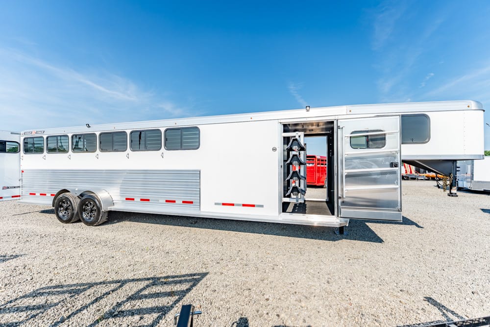 Long white aluminum horse trailer with an open side door, showing saddle racks inside on a gravel lot under a blue sky.