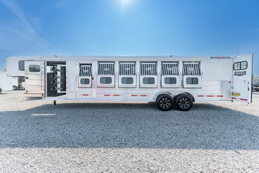Long silver horse trailer with multiple stall doors and barred windows parked on a gravel lot under a bright blue sky; Logan Coach branding visible on the rear.