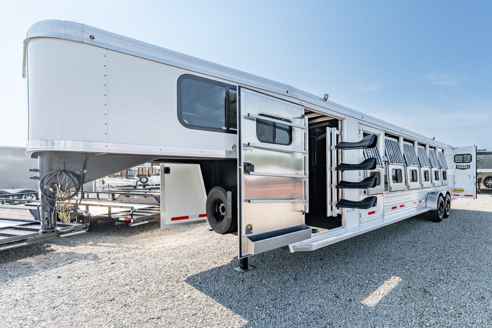 Long white horse trailer with multiple side stalls and an open entry door on a gravel lot under a clear blue sky.