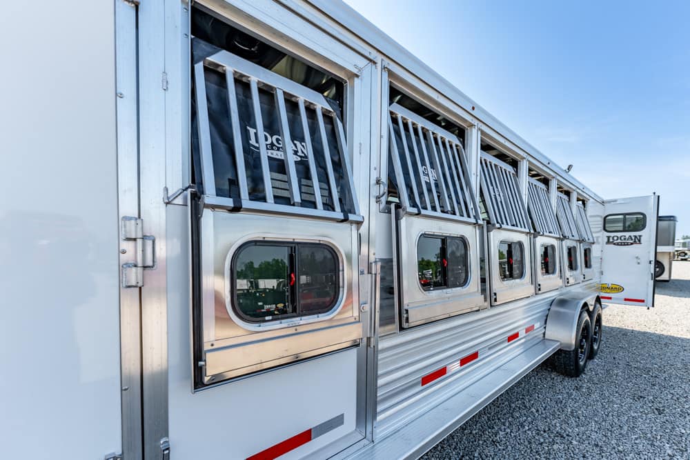 Aluminum horse trailer with multiple stall windows, doors open, on a gravel lot under a clear blue sky.