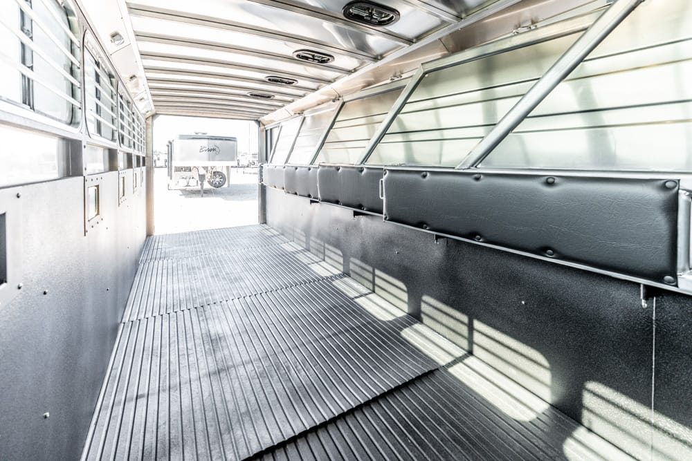 Interior of a metal horse trailer with padded side walls and ribbed floor, door open to bright outdoor light outside the trailer