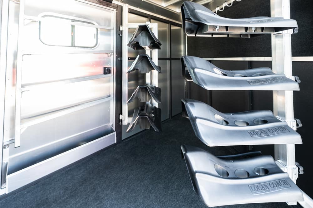 Interior of a horse trailer: stacked saddle racks on the right and a metal door on the left, with a dark carpeted floor.