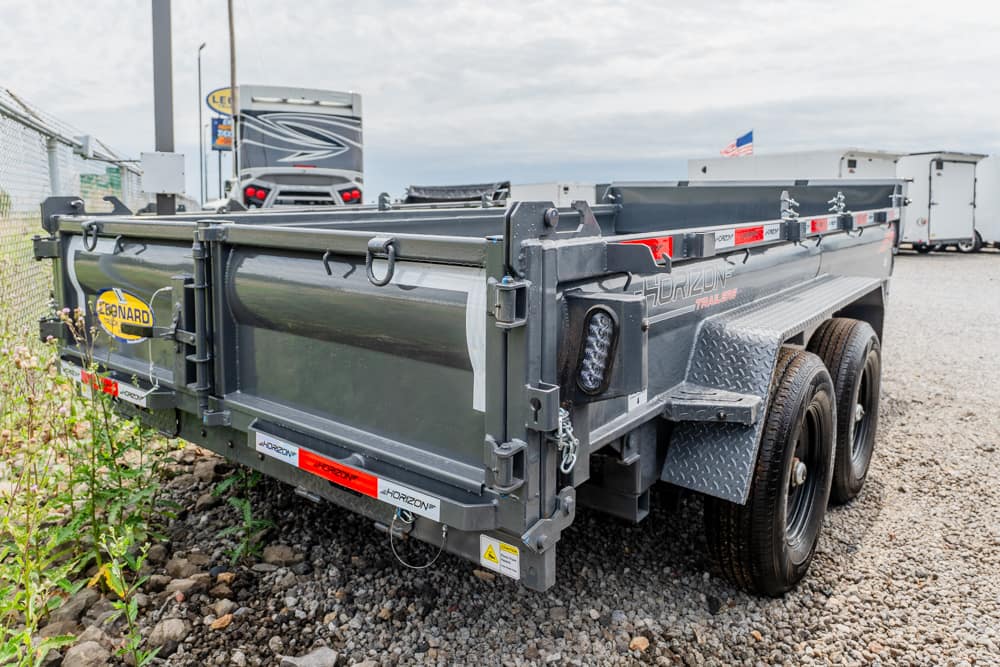 Grey Horizon dump trailer with dual axles on gravel, rear-left view showing latches and hooks