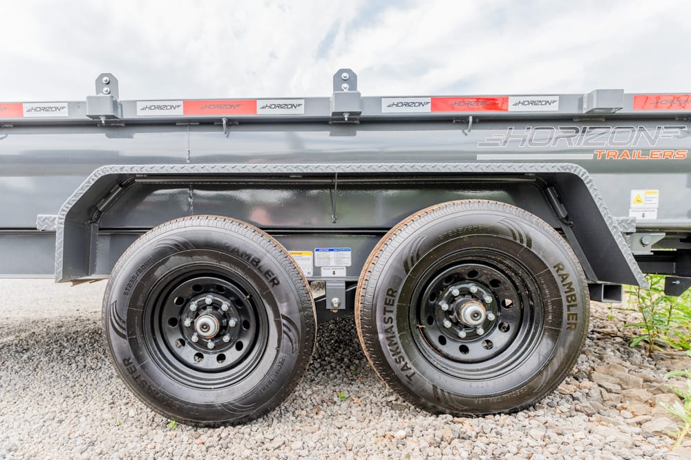 Dual-axle trailer with two black steel wheels on a gravel surface; tires labeled Rambler under a gray Horizon trailers body.