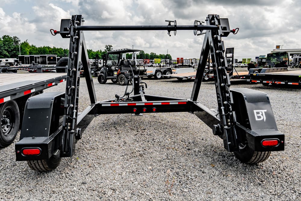 Heavy-duty equipment trailer frame in a gravel lot, with BT-branded fenders and dual ramp-like arms raised for transport.