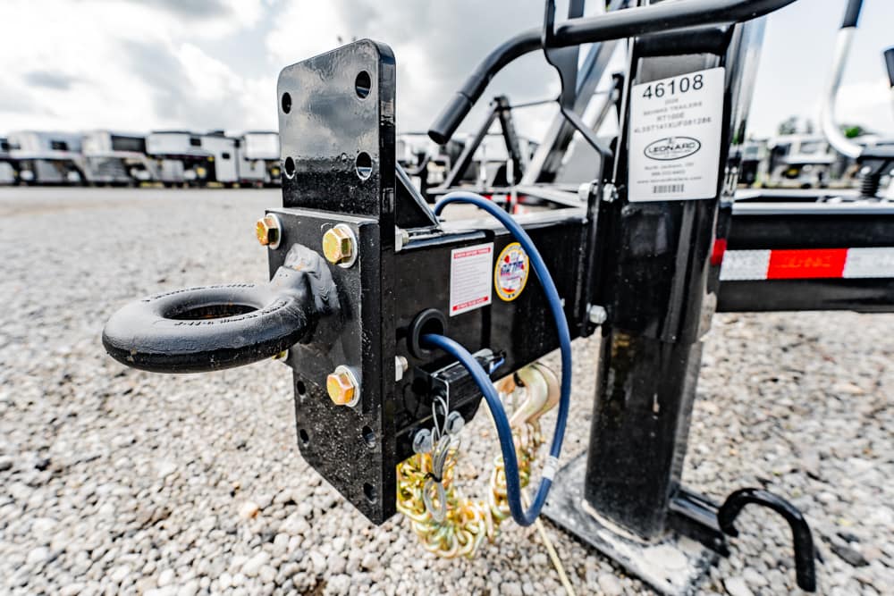 Close-up of a trailer hitch coupling with bolts, blue hoses, and safety chain on a gravel lot, trailers visible in the background.