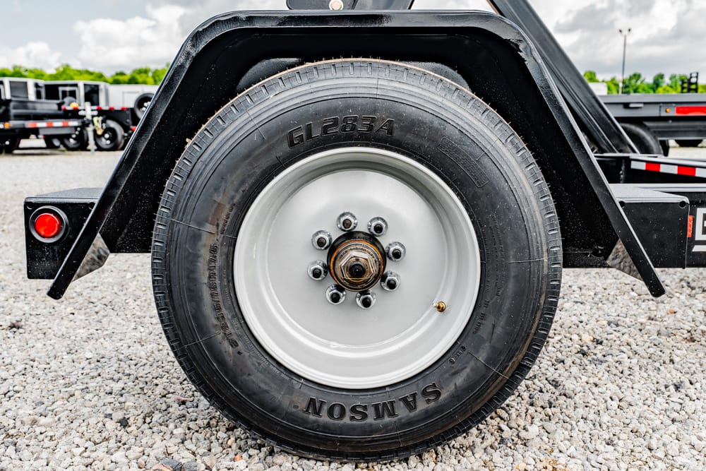 Close-up of a trailer wheel with black tire and silver rim on gravel, showing lug nuts and hub in focus