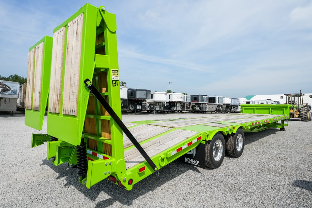 Bright green lowboy trailer with hinged rear loading ramps raised, parked on a gravel lot.